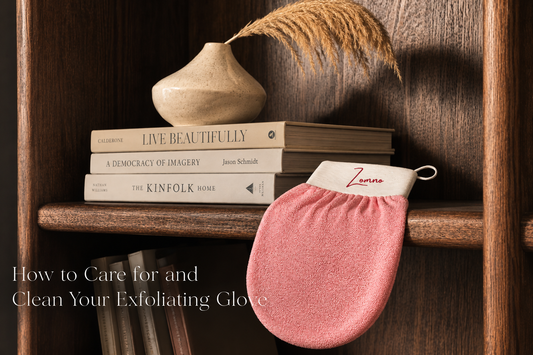 Zomno™ exfoliating glove draped over the corner of a wooden bookshelf next to a stack of neutral-toned hardcover books and a small ceramic vase with a single dried pampas grass stem. 