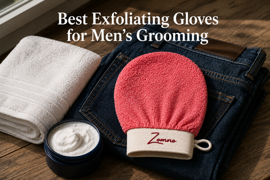A Zomno™ exfoliating glove resting on a folded pair of dark blue denim jeans and a simple white towel, a small pot of shaving cream beside it